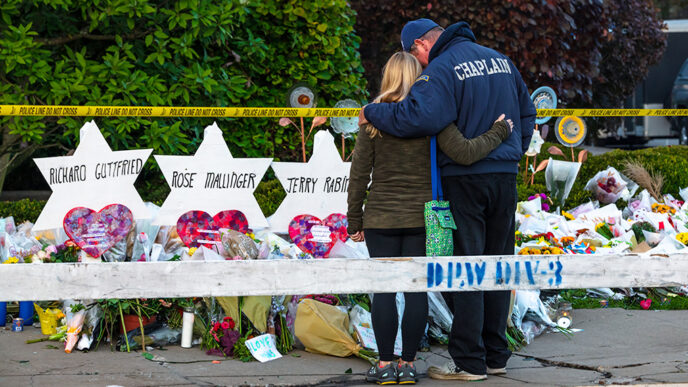 A man wearing a jacket that reads Chaplain embraces a woman as they stand in front of a row of memorials on a sidewalk|I Gave to #LoveIsStronger|The Pittsburgh Steelers team logo reworked to include a Star of David.