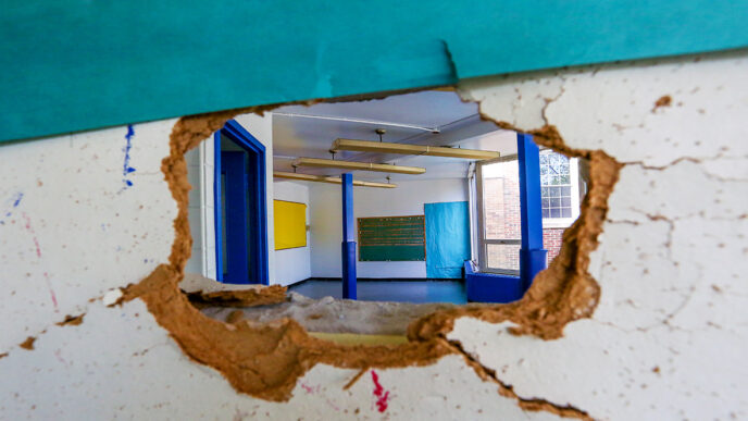 A hole in an interior wall shows a view into an empty room beyond|A woman stands with arms crossed.