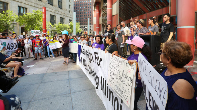 Speakers hold signs at a rally on the plaza surrounding a municipal building|A rally attendee holds up a sign reading I Am My Sister's Keeper|A group of women hold colorful hand-lettered signs in English and Spanish in support of DACA|Viewed from behind.