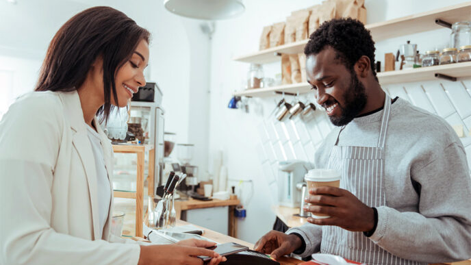 A smiling woman uses her phone to make a purchase at a coffee shop from an employee wearing an apron.