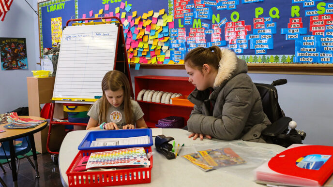 A teacher watches a student reading at a table in a brightly decorated classroom|A group of young children sit cross-legged on the floor facing a person using a wheelchair.