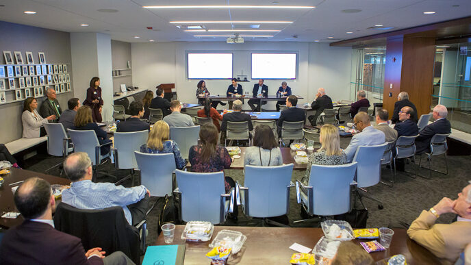 Four people seated at the front of a conference room face an audience in rows of chairs|A row of seated people.