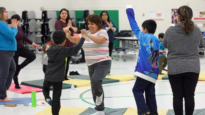 A group of adults and children stand in a yoga pose on mats in a gymnasium|A young boy kneels on a gym mat.