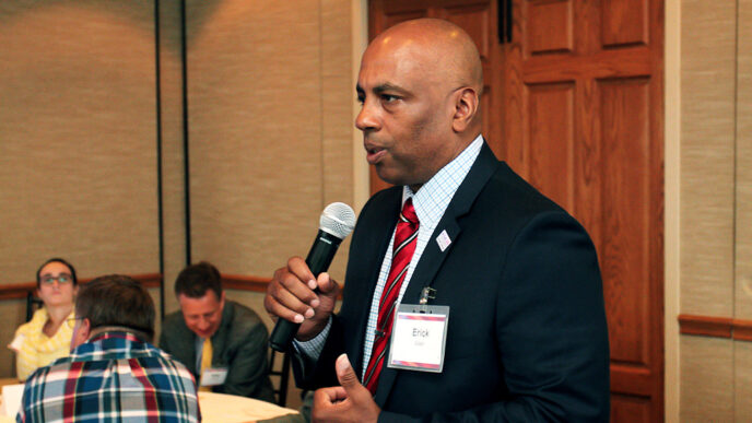 A man holding a microphone speaks to people seated in a conference room.