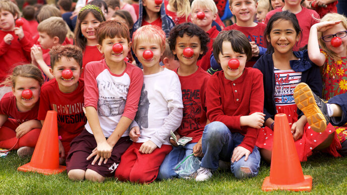 Rows of children sit on the sidelines of a grass playing field.