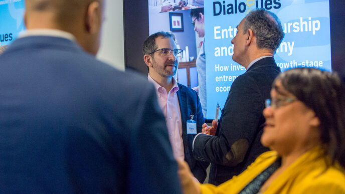 Two pairs of people in conversation standing in front of a projection screen|A row of people seated at a conference table lean to see and listen to a speaker|Around a conference table.