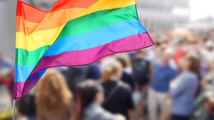 A rainbow flag flies in front of a crowd of people in soft focus.