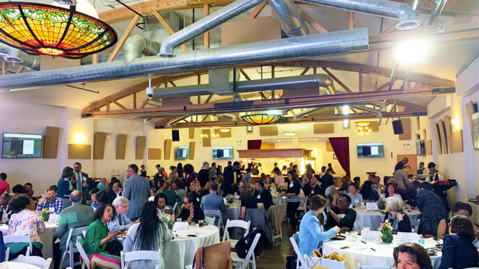 A crowd gathers around tables in in a room with high ceilings and stained glass chandeliers|African American Legacy logo|Asian Giving Circle logo|Nuestro Futuro logo|Young Leaders Fund logo.