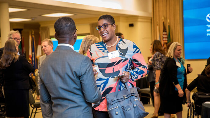 Two people standing and talking in a crowded meeting room|Three panelists seated on a stage in front of banners reading Disability Inclusion Opportunity Summit.