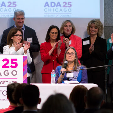 A group of people gathered behind a podium with a sign reading ADA 25 Chicago as one women speaks into a microphone|A group of people gathered behind a podium with a sign reading ADA 25 Chicago as one women speaks into a microphone.