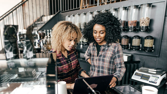 Two women behind the counter at a coffee shop look at a laptop.