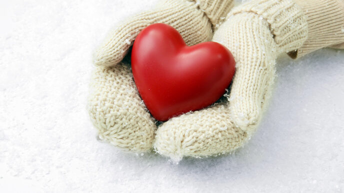 A pair of hands wearing mittens hold a red heart against a background of snow.